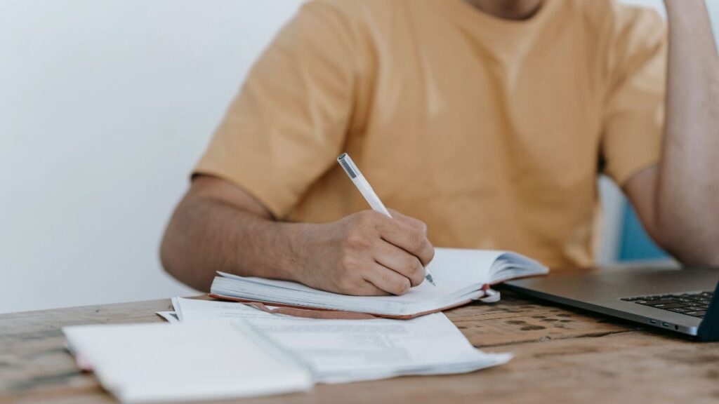Hombre joven sentado en un escritorio escribiendo sus objetivos financieros y gastos diarios en una libreta de papel.
